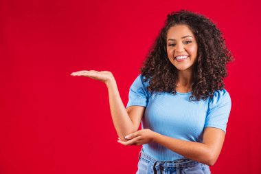 Neşeli Afro kadın fotokopi odasını işaret ediyor, inanılmaz reklamları tartışıyor, yol gösteriyor ya da yön veriyor, sarı kalın kazak giyiyor, hoş bir gülümsemesi var, iyimser hissediyor, kırmızı arka planda izole edilmiş..