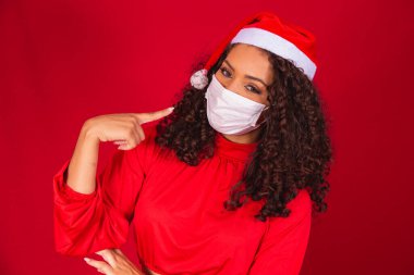 Noel şapkalı genç kadın Coronavirus virüsü virüsünden korunmak için işaret parmağıyla kırmızı arka plan stüdyosuna işaret ediyor. Mutlu yeni yıl kutlamaları, mutlu tatiller.