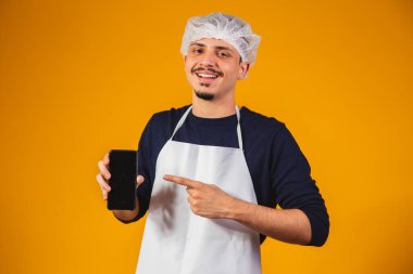 Önlüklü çocuk cep telefonunu işaret ediyor. Sarı arka planda gastronomi öğrencisi. Elinde üniforma ve cep telefonu var.