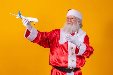 Noel Baba sarı arka planda küçük bir oyuncak uçak tutuyor. Noel yolculuğu konsepti
