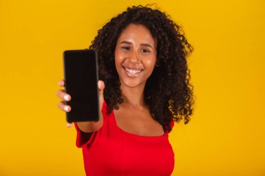 Noel şapkalı gülümseyen genç bir Noel Baba kadın. Elinde cep telefonu ve sarı arka planda boş bir ekran var..