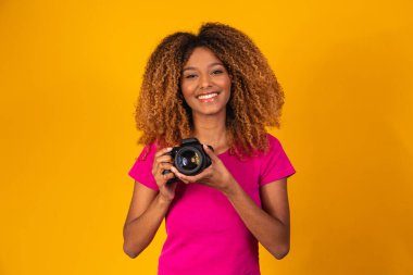 Genç afro fotoğrafçı elinde sarı arka planda bir kamera tutuyor..