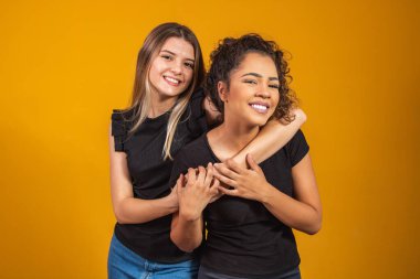Sarı arka planda, çeşitlilik kavramında birbirlerine destek olan farklı etnik kökenlerden arkadaşlar. Bir afro kadın ve bir sarışın birbirlerine sarılıp gülümsüyorlar kameraya bakıyorlar.