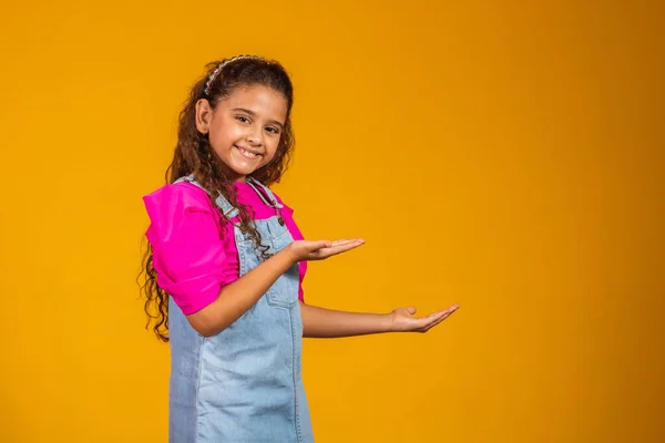 Stock image Positive beautiful mixed little girl on yellow background with beaming smile pointing to the side on empty copy space. Advertising concept.