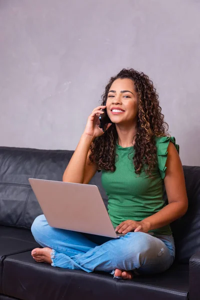 微笑的千禧年非洲裔美国妇女用手机在家庭办公室使用笔记本电脑 快乐的年轻混血女士打电话有手机对话看电脑屏幕坐在坐在屏幕上 — 图库照片