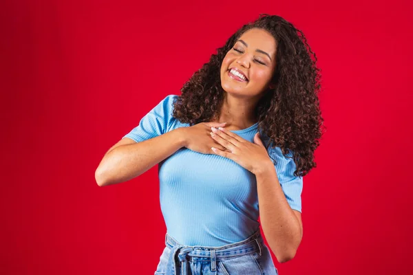 Grateful hopeful happy black woman holding hands on chest feeling pleased thankful, sincere african lady expressing heartfelt love appreciation gratitude honesty isolated on red studio background