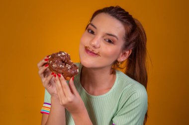 Güzel kızıl saçlı kız sarı arka planda çikolatalı donut yiyor. Kız donut yiyor.