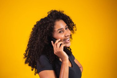 Sarı arka planda cep telefonuyla konuşan mutlu Afro-Amerikan genç kadın portresi.