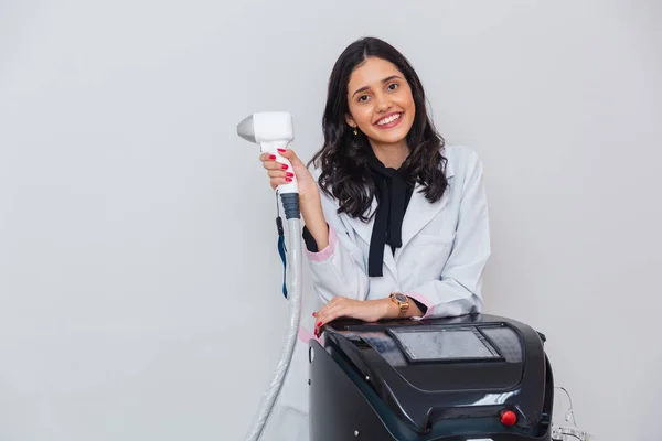 Brezilyalı, beyaz önlük giyen, vücut ve yüz kıllarını aldırmak için lazer tutan, lazer saçlı bir kadın. Tabanca ve makineyle.