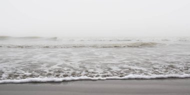 Sisli bir sabahta okyanus dalgaları kumlu deniz sahillerinde gri renkli köpükler yaratıyor..