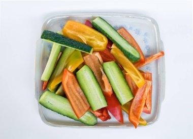 Beyaz arkaplanda kare şeklinde bir porselen tabakta sarı, kırmızı biber ve salatalık dilimlerinin üst görüntüsü.