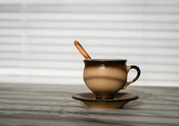 A brown ceramic cup with a ceramic spoon on a saucer standing on a stone tabletop and illuminated by the sun's rays through the blinds on the window