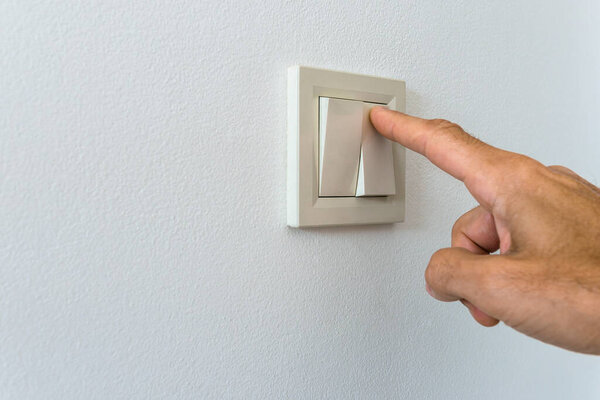 A man's hand presses the electric switch with his index finger on a white wall. Background