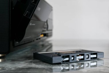 Bir kaset, cilalanmış mermer bir rafta, açık kasetçalar ve otomatik ters kaset kaydedicinin yanında duruyor..