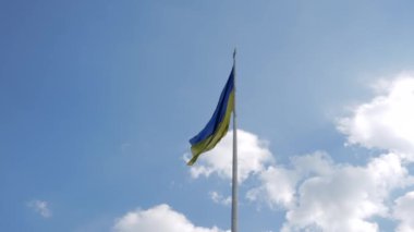 Ukrayna bayrağı yavaşça bir bayrak direğinde dalgalanıyor. Arkasında armalar ve bulutlar var.