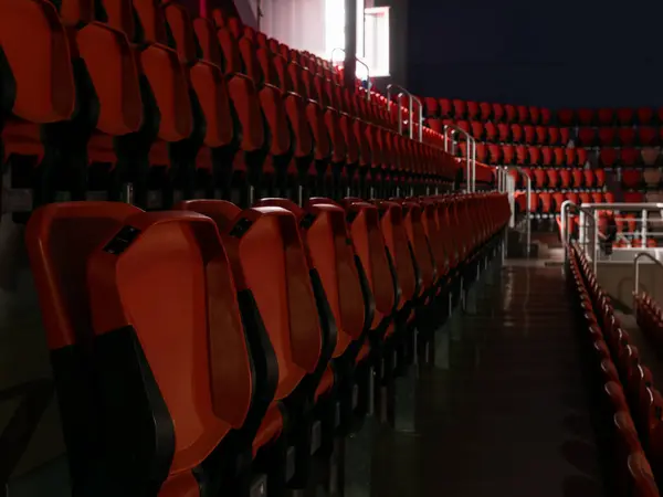 Koyu renkli bir spor salonunun tribünlerindeki kırmızı koltuklar sadece pencerelerden gelen ışıkla aydınlatılıyor.