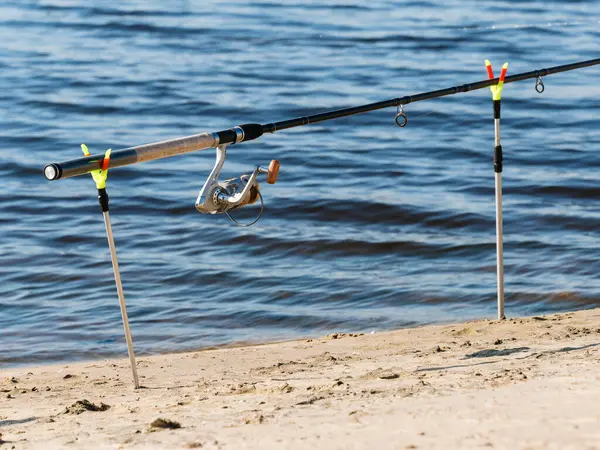 Kumlu bir sahil ve suyun arka planında iki tutucu üzerinde dönen balık oltası.