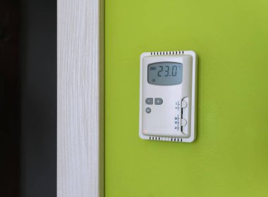 Parlak yeşil bir duvara monte edilmiş, termometresi 23 santigrat derece olan, duvara monte edilmiş bir klima kontrol paneli.