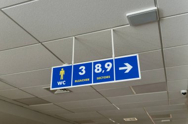 Halka açık bir alanda tavanda asılı duran mavi bir tabela tuvaletleri, stantları ve oturma alanlarını gösteriyor..