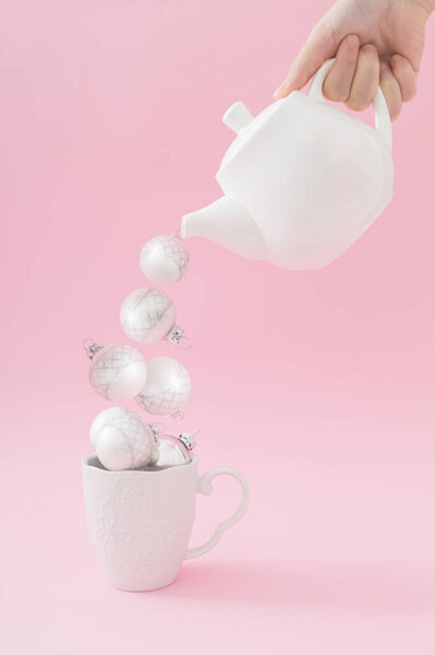Creative composition with hand holding teapot and pouring Christmas baubles into tea cup on pastel pink background. Minimal Christmas or New Year concept. Trendy winter holidays idea.