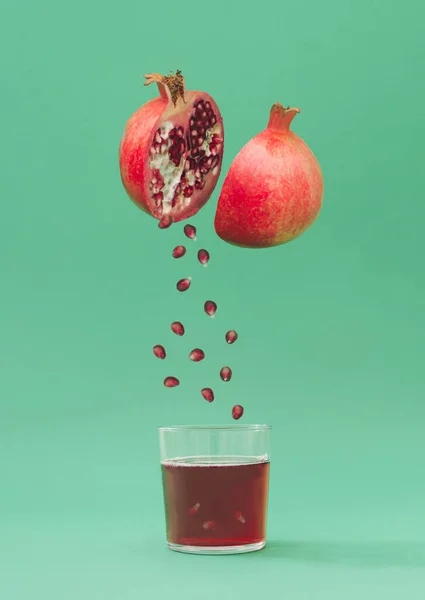 Creative layout of pomegranate and pomegranate seeds falling into transparent glass full of red pomegranate juice on pastel green background. Minimal fruit concept. Trendy healthy food idea.