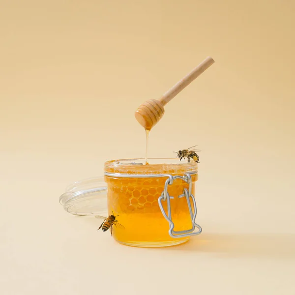 Creative layout made with honey dripping from honey dipper in jar full of natural honey and honeycombs and honey bees flying around on light beige background. Minimal nature concept. Trendy food idea.