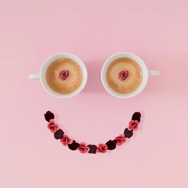 Layout of smiley emoticon made with coffee cups and flowers on pink background. Minimal coffee concept. Creative positive thinking and good mood idea. Coffee and flowers aesthetic. Flat lay.