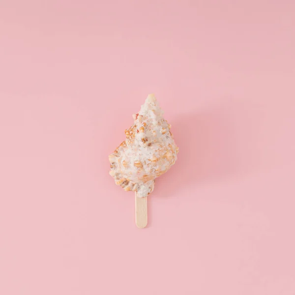 Trendy summer layout of sea shell with ice cream stick on light peachy pink background. Minimal summer concept. Creative beach food idea. Ice cream aesthetic. Flat lay. Top of view.