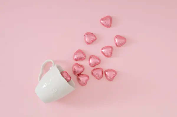 Beyaz kahve fincanından gelen pembe folyoya sarılı çikolata kalpleriyle yapılmış yaratıcı bir aşk kompozisyonu. Asgari aşk konsepti. Romantik çikolata kalp fikri.