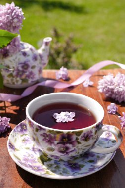 Üzerinde çiçekler olan mor bir kupa, çaydanlık ve açık arkaplanda leylak çiçekleri olan bahar kompozisyonu. Kapatın. Çay içiyorum. Menü, tebrik kartı. Bahar geldi. 'Günaydın' konsepti 
