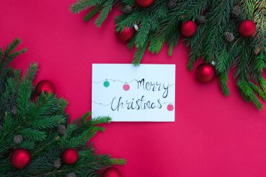 Renkli kartpostallı Noel kompozisyonunun en üst görüntüsü Mutlu Noeller yazılı. Boş tebrik kartı, köknar dalları ve kırmızı arka planda topları olan davetiye modeli. Seçici odak.