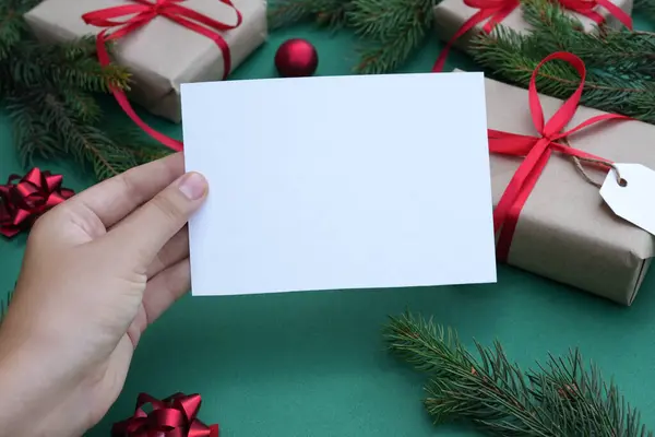 Blank paperin female hands. Flat lay of green background with gift boxes, Christmas decoration and fir tree branches. Top view mock up and copy space for text. Christmas background.