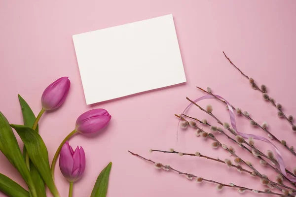 Mother\'s Day or International Womens-day concept. Top view of white paper blank, fresh tulips and pussy willow branches on pink background. Flat lay. Mock up. Space for text.