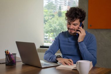 Genç, gülümseyen, serbest çalışan bir Latin 'in portresi. Bilgisayarla çalışırken telefonla konuşuyor ve notlar alıyor.