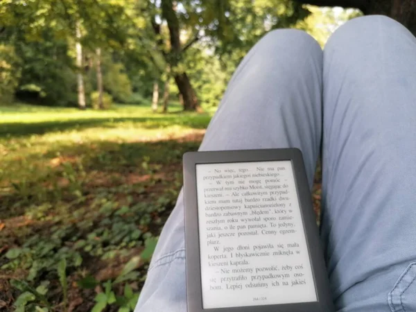 一个在公园里看书的人 膝盖上放着一个电子阅读器 绿树成荫 绿草成荫 宁静祥和 — 图库照片