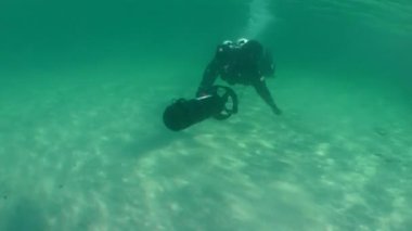 Su altı scooter 'ı olan bir dalgıç sığ sularda kumlu bir zeminde yüzer, sonra kamera kıyı kentsel manzarasını göstermek için suyun üzerinde döner..