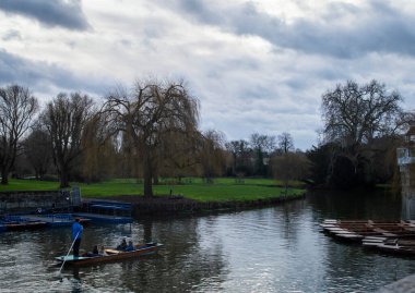 Cambridge, İngiltere, 7 Ocak 2022. Cambridge şehrinde bulutlu bir günde etrafı ağaçlar ve yeşil çimlerle çevrili ziyaretçilerle dolu bir tekne manzarası.. 