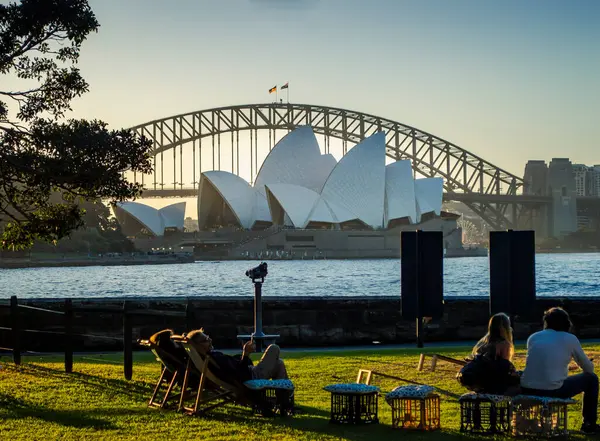 Sydney, Avustralya, 26 Temmuz 2023. Sydney Opera Evi 'nin rıhtım manzarasında gün batımından önce Royal Botanic Garden' da dinlenen insanlar var.. 