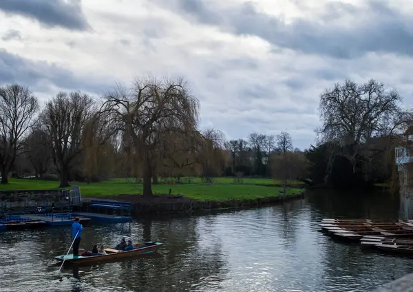 Cambridge, İngiltere, 7 Ocak 2022. Cambridge şehrinde bulutlu bir günde etrafı ağaçlar ve yeşil çimlerle çevrili ziyaretçilerle dolu bir tekne manzarası.. 