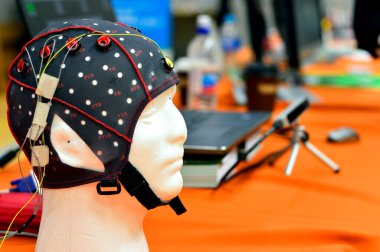 Elektroensefalogram (EEG) başlığı düz metal diskler (elektrotlar) ile bir bilim sergisinde gösterilen ve dizüstü bilgisayarların arka planda bulanıklaştığı beyaz plastik bir kafaya bağlıdır..