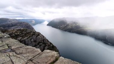 Norveç 'ten nefes kesici ikonik Preikestolen (Kürsü Kayası), sarp dağlar ve yağmurlu bir günde sisli bulutlarla çevrili dramatik bir fiyorda bakıyor. Sahne 4K yatay görüntüler Norveç doğasının güzelliğini gösteriyor. 