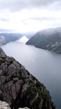 Norveç 'in Preikestolen (Minber Kayası) kıyısından dramatik POV görüntüleri, yağmurlu bir günde yüksek tepeler ve alçak asılı bulutlarla çevrili görkemli fiyortlara bakıyor. Dikey 4K görüntüler Norveç doğasının ham güzelliğini yansıtıyor.