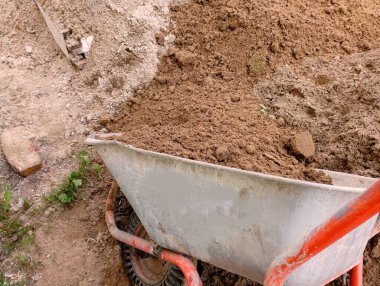 Bir inşaat el arabası yığına kum döker. İnşaat alanında el arabası kullanılarak toplu malzeme nakli. Turuncu saplı ve kumlu inşaat el arabası..