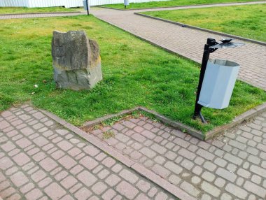 Parktaki kaldırımın kenarındaki çöp tenekesi. Çimlerin ortasında büyük bir taş. Şehir manzarası.