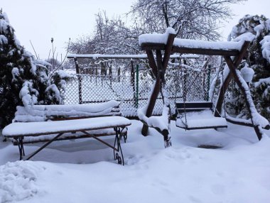 Bir bahçe masasının yanında kışın sokağın ortasında kurumuş meşe dallarından yapılmış ahşap bir salıncak kalın bir kar tabakasıyla kaplıdır.