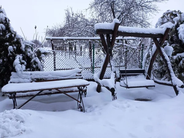 Bir bahçe masasının yanında kışın sokağın ortasında kurumuş meşe dallarından yapılmış ahşap bir salıncak kalın bir kar tabakasıyla kaplıdır.