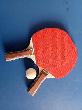 Two ping pong rackets lie on a blue tennis table. A white ball lies between the ping-pong rackets. Sports theme.