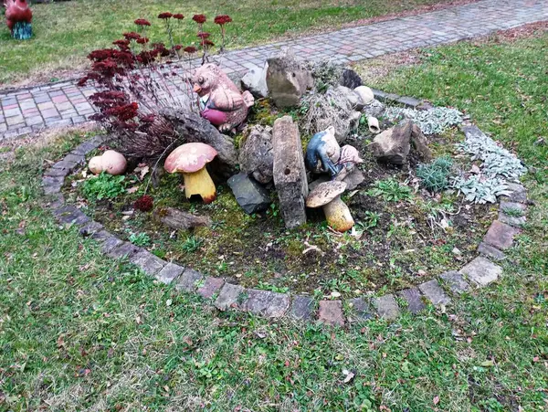 A round flowerbed is made of river stone, on which old plaster figures of animals are placed. Landscape design in the yard. Flowerbed with flowers and stones with figures of mushrooms.