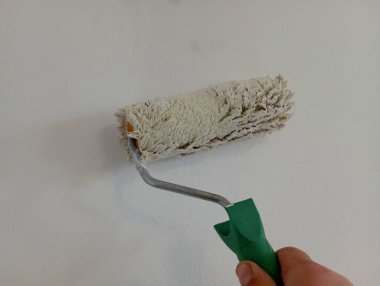 Duvarları, yeşil saplı bir silindirle beyaz renge boyamak. Fotoğrafta, bir insan eli elinde bir duvar boyası silindiri tutuyor ve odanın duvarlarını beyaza boyuyor. Ev onarımı konusu.