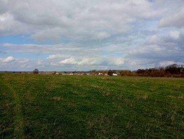 Ufkun ötesindeki manzarada küçük bir köy görülebilir. Sakin mavi gökyüzünün altında, üzerinde beyaz bulutlar yüzen güzel çimenli bir tarla..